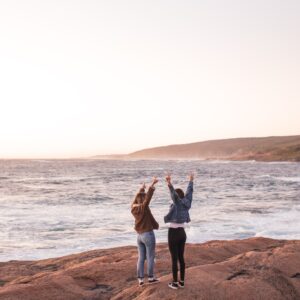 A photo of two people standing in front of the ocean, holding hands and raising their arms in the air. Located near Opus Health