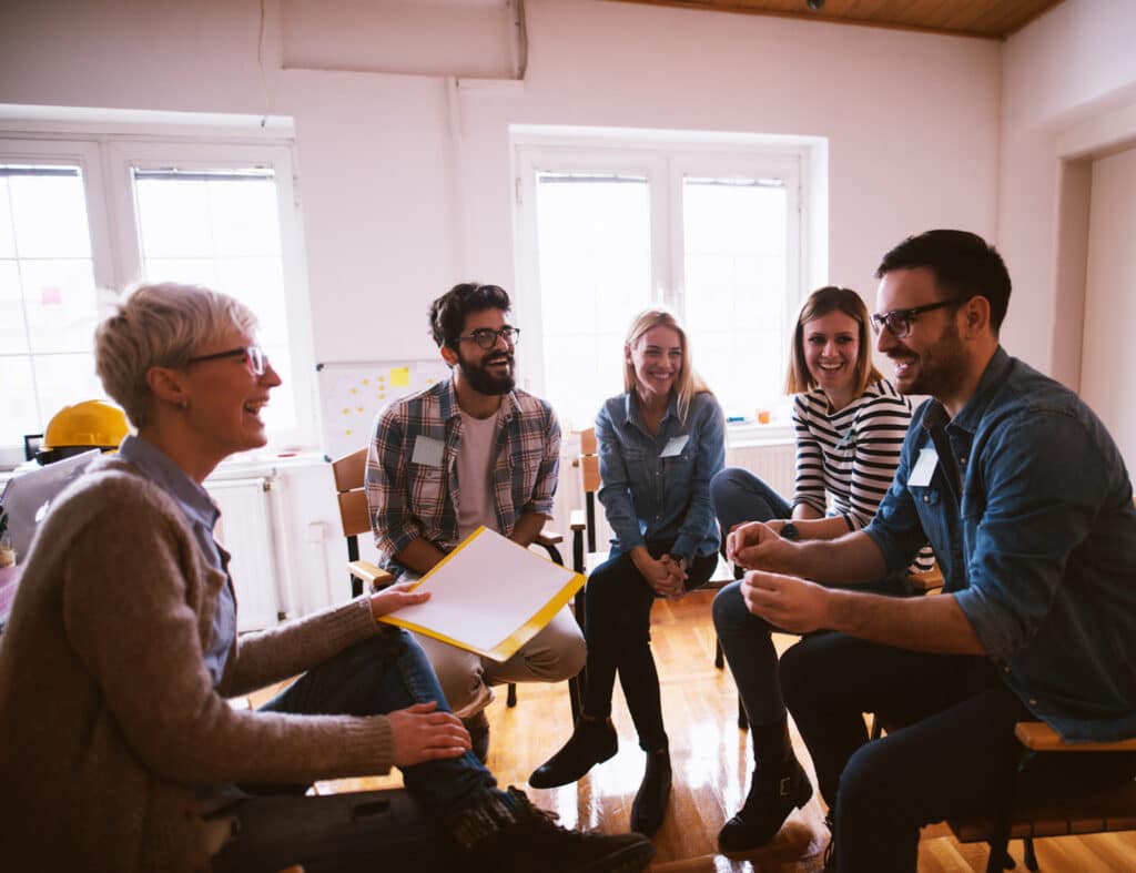 A photo of a group of people in group therapy, smiling and talking.-Support Groups Can Make a Difference in Addiction Recovery. Group of People in Group Therapy Feeling Positive.