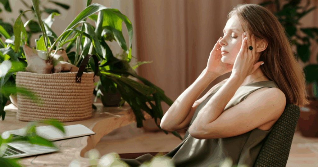 Woman experiencing Klonopin withdrawal symptoms, holding head in pain. Natural light, plants, laptop in background.