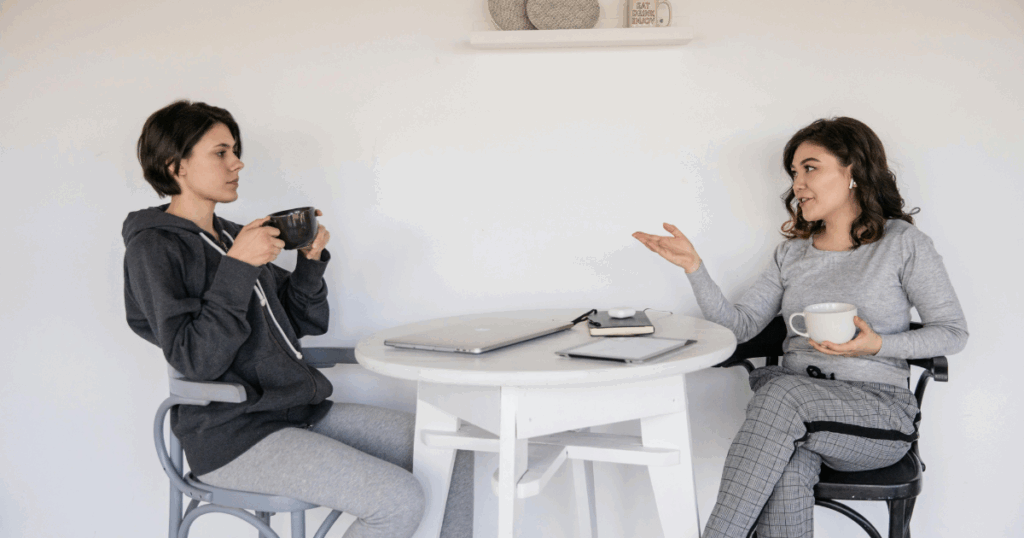 Two women discussing oxytocin withdrawal symptoms over coffee at a table with a laptop and tablet.