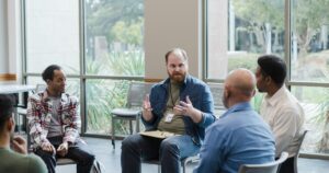 Five men sit in a circle in a bright room; the bearded man in the center speaks with hand gestures and wears an ID badge.