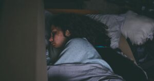 Person sleeping on a bed with a black cat cuddled against their back on striped sheets.
