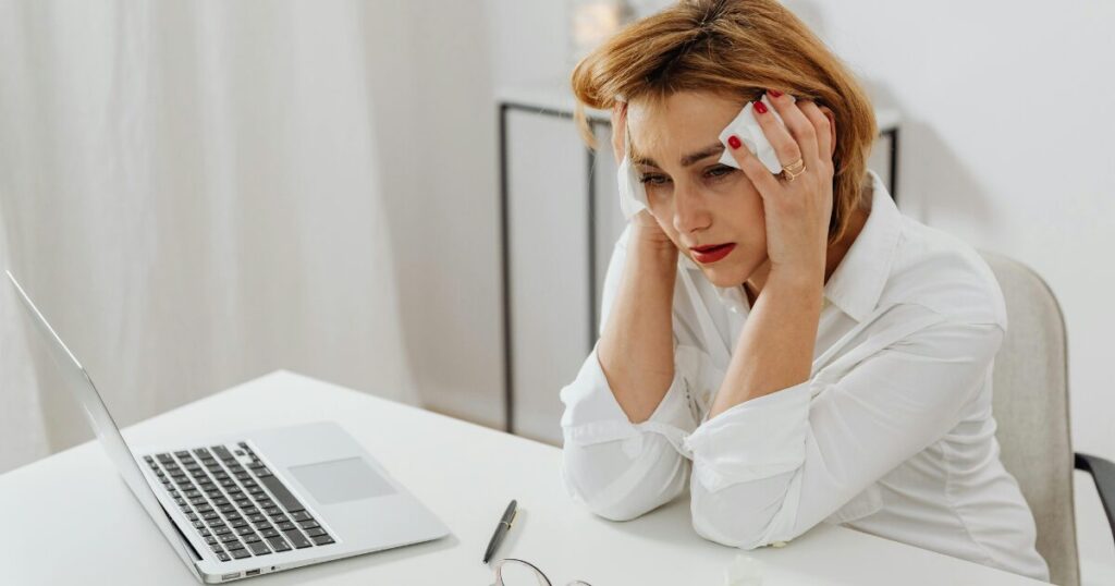 Woman in a white blouse sits at a desk with a laptop, holding tissues to her temples in distress.