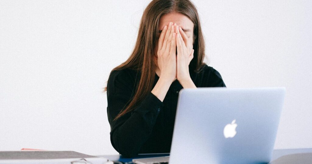 Woman sits at a desk with an open laptop, hands covering her face in distress.