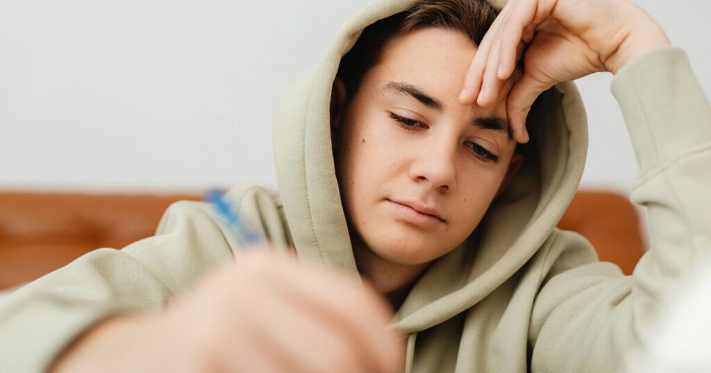 Young person in a beige hoodie rests their head on one hand, looking down at a card held in the other hand.