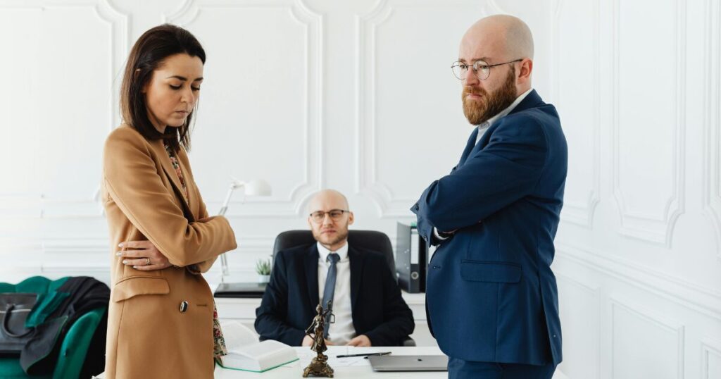 Two professionals stand with arms crossed in a bright office while a seated colleague watches from behind the desk, suggesting a tense discussion in progress.