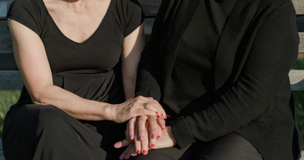 Two people sit closely on a bench, holding hands with rings visible on their fingers and painted nails in red and purple hues.