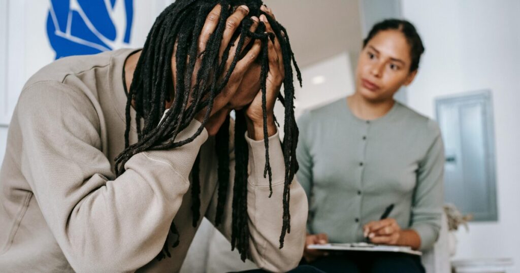 Person sits with head in hands, showing distress during a counseling session with a caregiver taking notes in the background.