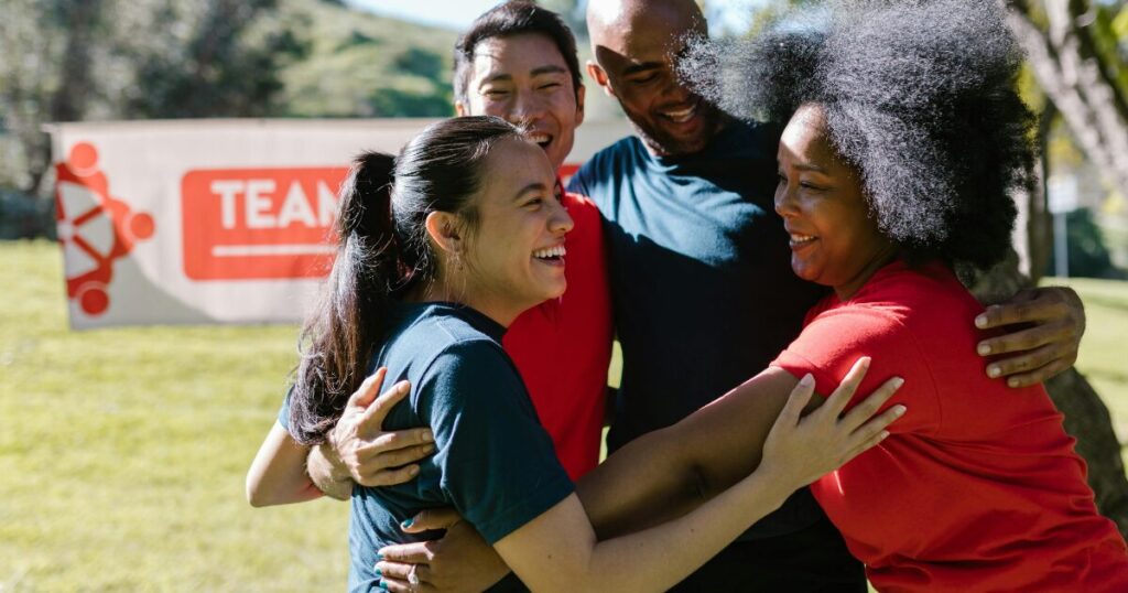 Group of four diverse friends hugging in a sunlit park with a TEAM banner in the background