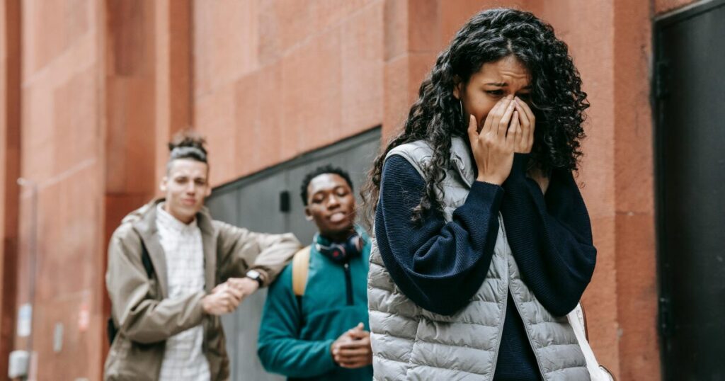 Young woman in a gray puffer vest covers her face in distress, while two friends stand in the background on a city street.