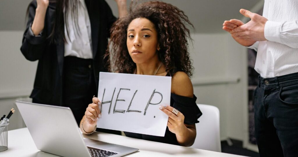 image - Opus Health Woman sitting at a desk holds a handwritten 'HELP' sign toward the camera beside a laptop, appearing distressed in an office setting, with colleagues nearby.