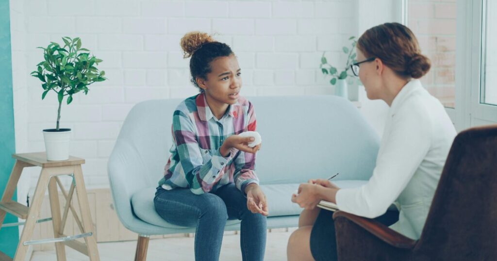 Teen girl on a light blue couch holds a stress ball and talks to a counselor taking notes in a bright, cozy room with plants behind them.