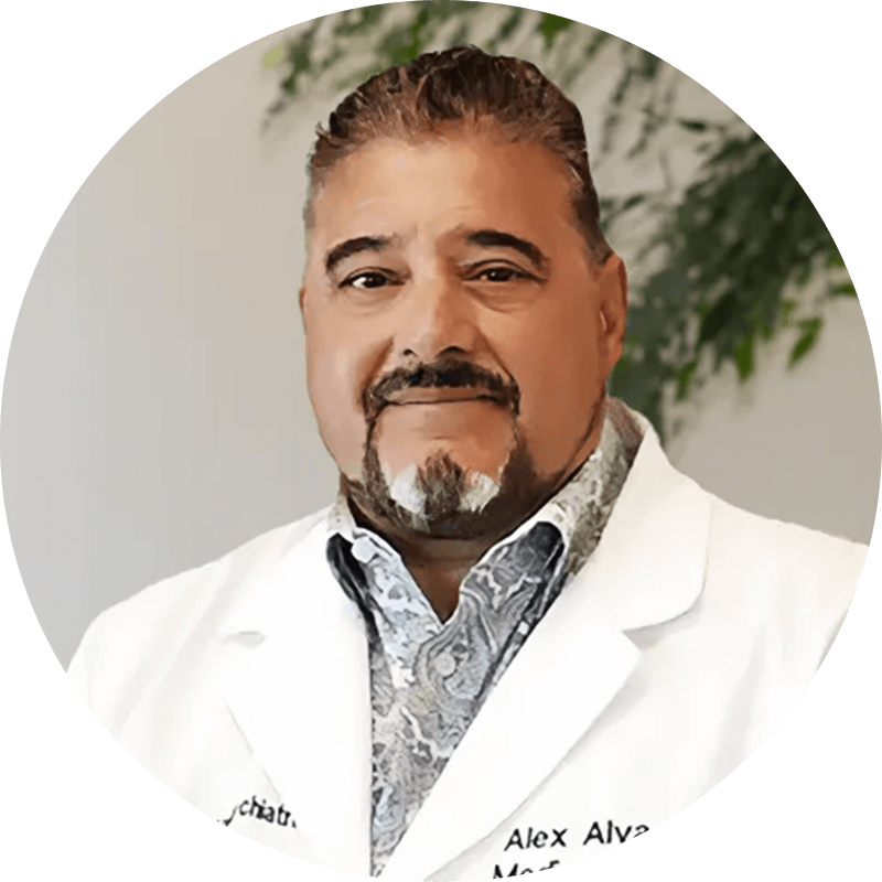 Dr-Alejandro-Alva-1-1png - Opus Health Portrait of a smiling male doctor in a white coat, with a patterned shirt, standing indoors with greenery in the background