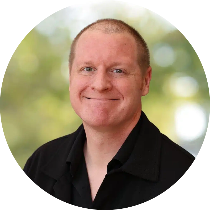 image-17webp - Opus Health Smiling man in a black collared shirt, studio-style portrait with a blurred green background