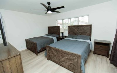 LP Facilities Tour: Bedroom with two beds, wood frames, gray bedding, ceiling fan, and natural light from window.