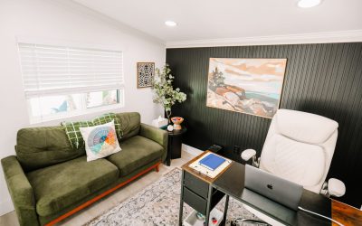Modern home office interior with green sofa, desk with laptop, and coastal painting, creating a cozy workspace.