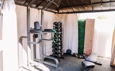 Home gym setup with weight rack, pull-up station, adjustable bench, kettlebells, and medicine ball in covered outdoor space.
