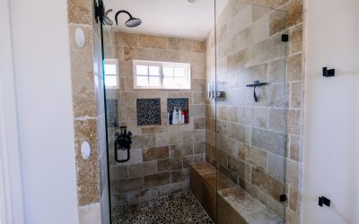 Walk-in shower with travertine tile, pebble floor, glass door, shower bench, and black fixtures in a modern bathroom.