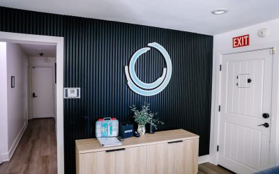 Modern office reception area with a blue circular logo on a black slat wall, light wood cabinet, and an exit door.
