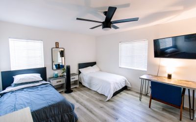 Modern bedroom with two twin beds, desk, TV, and ceiling fan. Interior design with white walls and wood-look flooring.