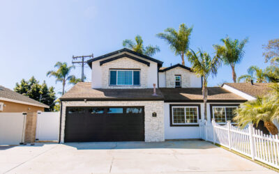 Exterior of a two-story house with palm trees, white stone facade, black garage door, and white picket fence.